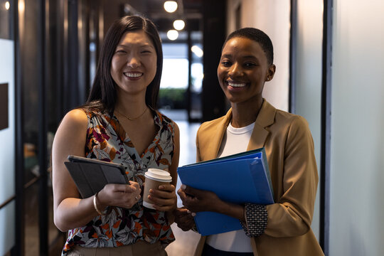 Portrait of happy diverse female colleagues with coffees, tablet and paperwork smiling in corridor