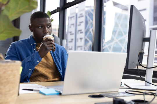 Thoughtful african american casual businessman drinking coffee and using computer at desk in office