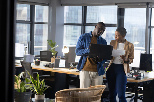 African american male and female colleague using laptop and talking standing in office, copy space