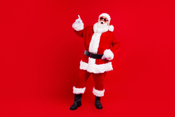 Portrait of ecstatic funny santa claus with white beard in sunglass directing at billboard empty space isolated on red color background