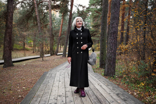 Graceful mature woman dressed in chic black coat, stroll through enchanting forest, reveling in splendor of nature with serene smile, grace and relaxation