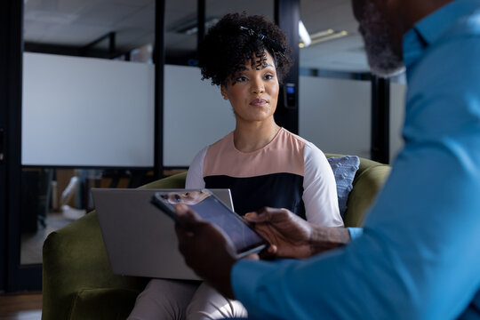 Focused diverse female and male colleague using laptop and tablet talking in office lounge