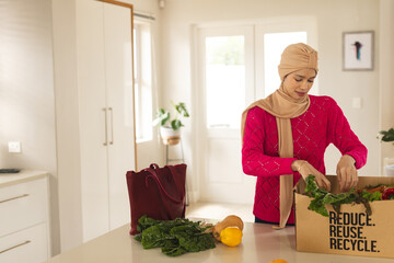 Biracial woman in hijab unpacking groceries from cardboard box in kitchen