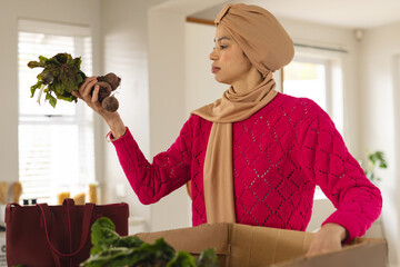 Biracial woman in hijab unpacking groceries from cardboard box in kitchen