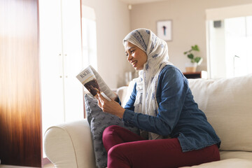 Happy biracial woman in hijab reading book in living room