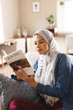 Biracial woman in hijab reading book in living room