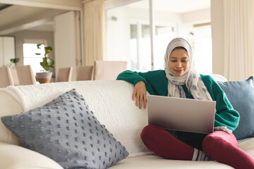 Happy biracial woman in hijab using laptop in living room