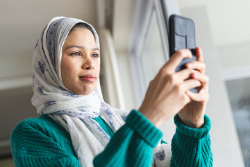 Biracial woman in hijab using smartphone at sunny home