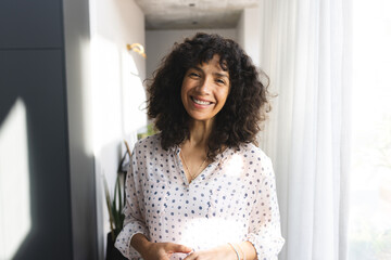 Portrait of happy caucasian mature woman with long curly dark hair, smiling in sunny room at home