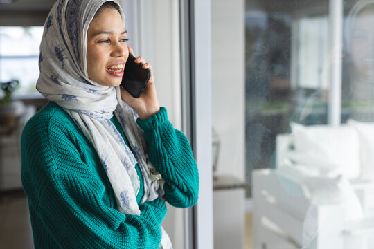 Happy biracial woman in hijab talking on smartphone at sunny home - Powered by Adobe