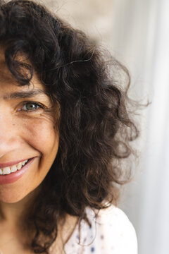 Half portrait of happy caucasian mature woman with long curly dark hair, smiling in sunny at home
