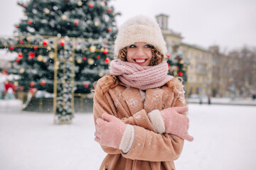 A beautiful curly girl in a snow-covered coat is a little cold. Happy woman posing against the background of a New Year tree in the park on a beautiful December day.