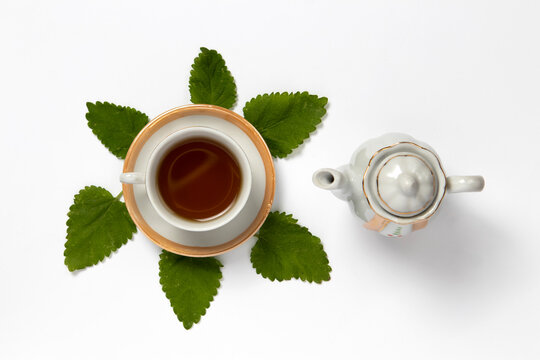 Top view of a tea in a teacup with fresh lemon balm leaves and a teapot on a white background