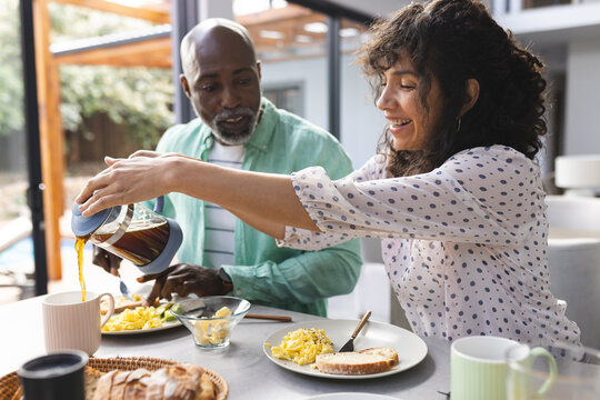 Happy mature diverse couple having breakfast and pouring coffee in sunny kitchen