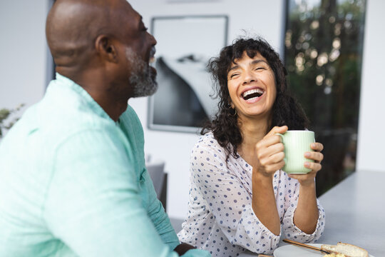 Happy mature diverse couple having coffee and laughing sitting in kitchen