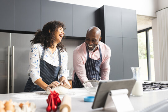 Happy mature diverse couple baking and using tablet in sunny kitchen, copy space - Powered by Adobe