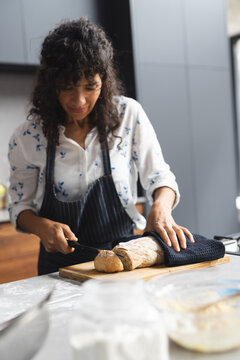 Happy mature caucasian woman cutting freshly baked bread in sunny kitchen, copy space