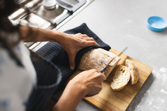 High angle of mature caucasian woman cutting fresh bread on breadboard in sunny kitchen, copy space