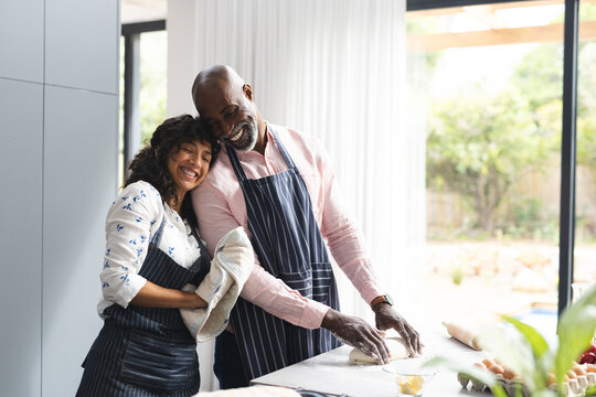 Happy diverse couple preparing dough and embracing, baking in sunny kitchen, copy space