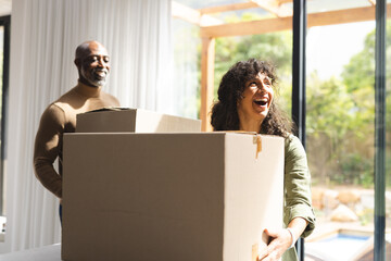 Happy diverse mature couple carrying packing boxes into sunny new home