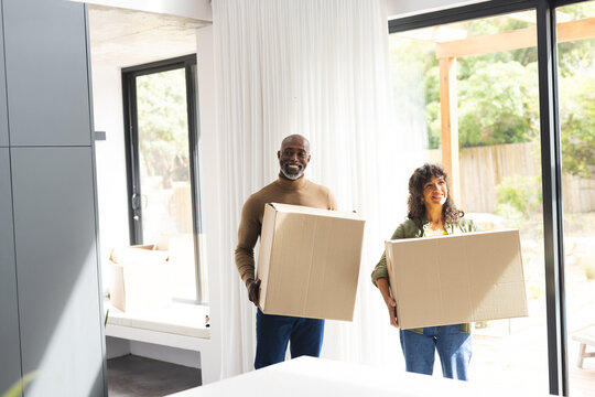 Happy diverse mature couple carrying packing boxes into sunny new home, copy space
