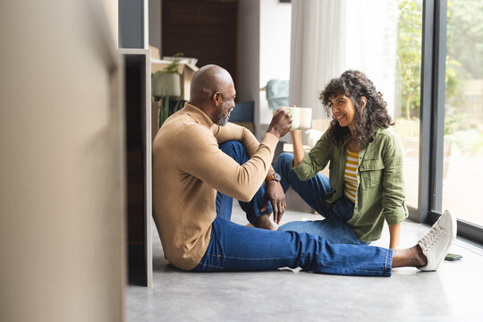 Happy diverse mature couple on floor toasting with coffees in sunny new home, copy space