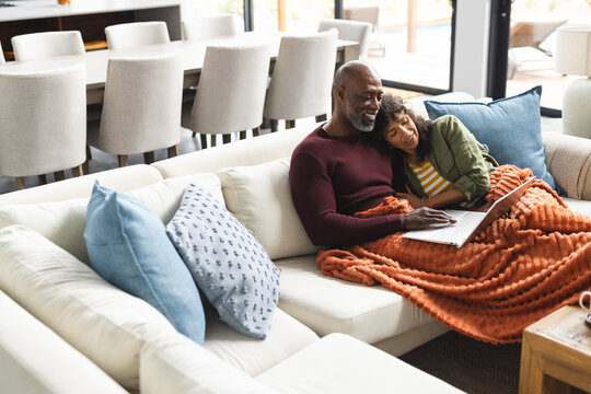 Diverse mature couple embracing on couch with blanket using laptop in living room, copy space