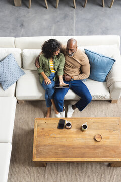 Happy diverse mature couple sitting on couch using tablet in living room, copy space