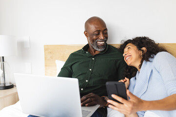 Happy diverse mature couple sitting on bed using laptop and smartphone in bedroom