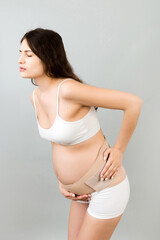 Portrait of pregnant woman in underwear wearing maternity belt on her belly and showing thumb up gesture at gray background. Orthopedic abdominal support belt concept. Copy space