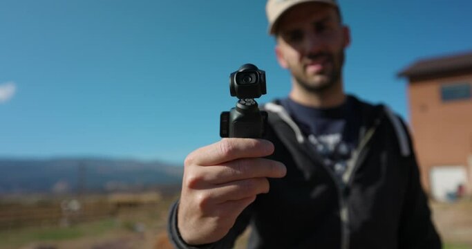Male Freelancer Holding 3-Axis Stabilized 4K Handheld Camera And Moving Around With Hand. Bokeh Background With Blue Sky. Slow Motion
