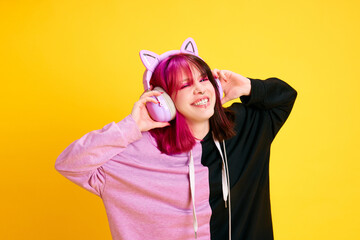 Happy, smiling young girl with piercing and two colored hair listening to music in headphones over vivid yellow studio background. Concept of youth, self-expression, fashion, emotions, positive mood