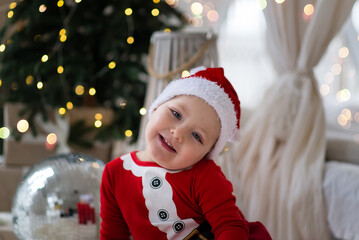 Smiling little boy in a red suit and Santa hat sitting near the Christmas tree, celebrating the New Year at home