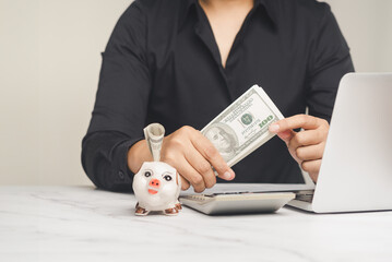 A piggy bank with a $100 bill with a background of a woman holding US dollar bills while sitting at a desk