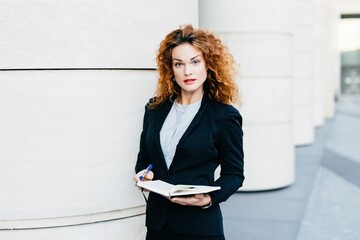 Focused businesswoman with curly hair writing in a notebook, poised against a sleek building...