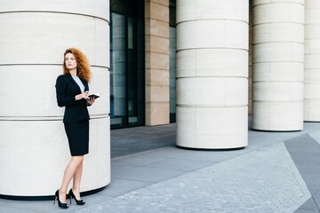 Solemn businesswoman with curly hair, holding a notebook, stands by architectural columns,...