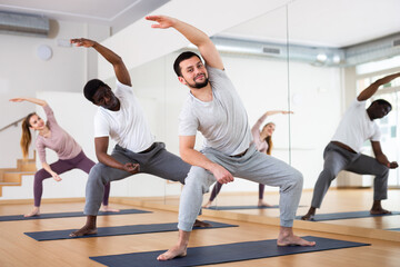 Group of people doing stretching exercises in squatting position on mats in fitness studio.