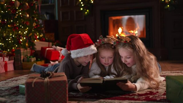 Merry Christmas Xmas holiday. Brother sisters children kids reading magic Christmas story book near Christmas tree in living room with fireplace in winter evening. Christmas eve at home