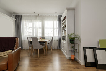 Living room of a house furnished with a circular dining table with chairs upholstered in gray fabric next to a large window with curtains