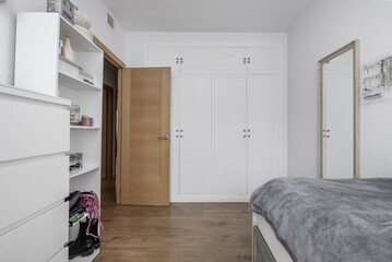A room in an apartment with a built-in wardrobe with white wooden doors with trunks in the attics, a wooden door and white furniture that matches the wardrobe