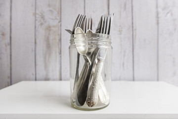 A clear glass jar filled with metal kitchen forks and knives on a white wooden cabinet