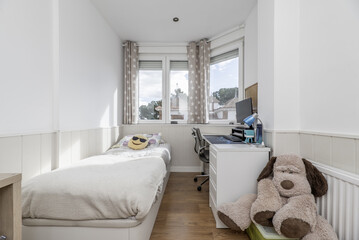 A youth bedroom with a desk under the aluminum window, white furniture, a bed with a canape, a large stuffed dog, matching walls with a wooden floor.