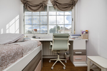 A beautiful youth room furnished in white and light tones with a desk under the window and wooden...