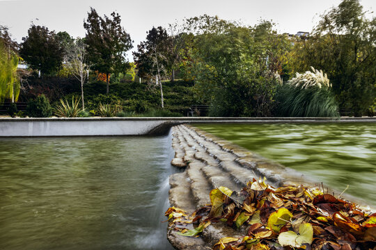 Long exposure image of small slope with falling water in a canal in an urban park with fallen leaves and lots of trees