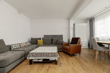 A spacious living room with a corner sofa upholstered in gray fabric, a square coffee table with a...