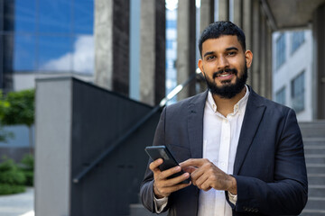 Close-up photo of a smiling young Arab man standing on the street near the office, holding a phone and smiling at the camera