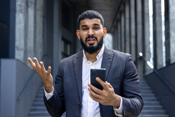 Close-up portrait of a shocked young Muslim businessman man holding a phone in his hands, standing outside on the street and throwing up his hands in frustration - obrazy, fototapety, plakaty