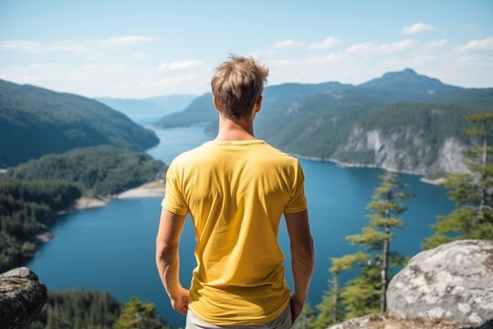 rear view of Young man in yellow t-shirt staying at the cliff looking at the fjord like lake and mountain viewpoint during sunny day, aesthetic look