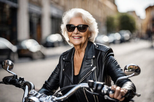 Happy beautiful elderly senior woman with grey hair rides a motorcycle along a city street, laughing and smiling, mature old biker in black leather jacket