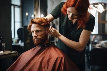 Gartenposter Stylish fashionable man with red hair and beard in a barbershop . Professional stylist woman barber, barber does styling for his client in a modern salon.  © Hope